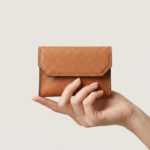 May include: A small, rectangular, tan-coloured wallet with a pebbled leather texture. The wallet features a fold-over flap closure with visible stitching along the edges. The design is compact and simple, showcasing a consistent grain pattern.