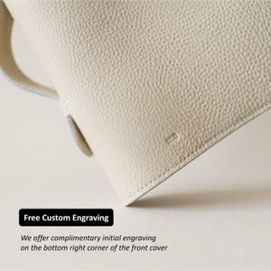 May include: Close-up of a cream-colored leather bag with a textured surface. The bag has a handle and a small, embossed "JEB" logo. The image includes the text "Free Custom Engraving".