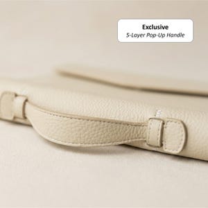 May include: Close-up of a cream-colored leather bag with a 5-layer pop-up handle. The handle is attached with small, matching leather loops. The bag has a textured surface and a minimalist design. The text "Exclusive" and "5-Layer Pop-Up Handle" is visible.