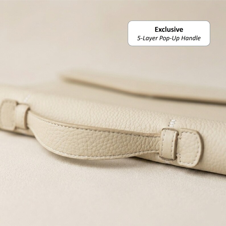 May include: Close-up of a cream-colored leather bag with a 5-layer pop-up handle. The handle is attached with small, matching leather loops. The bag has a textured surface and a minimalist design. The text "Exclusive" and "5-Layer Pop-Up Handle" is visible.