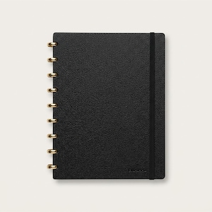 May include: A black notebook with a textured cover and gold-colored binding rings. A black elastic band secures the notebook. The notebook is rectangular and has a minimalist design.