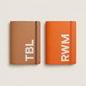 May include: Two rectangular notebooks with textured covers, one in tan and one in bright orange. Each notebook features a vertical elastic band closure and large white capital letters printed on the front, reading TBL on the tan and RWM on the orange.
