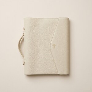 May include: Cream-colored Bible cover with a handle and a gold cross emblem. The cover has a textured surface and a stitched design on the front. The handle is attached with gold-colored hardware.