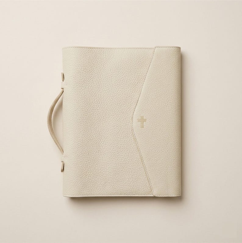 May include: Cream-colored Bible cover with a handle and a gold cross emblem. The cover has a textured surface and a stitched design on the front. The handle is attached with gold-colored hardware.