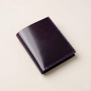 May include: A rectangular, bi-fold wallet crafted from smooth, deep purple leather. The wallet features a clean, minimalist design with visible stitching along the edges, showcasing a polished, reflective surface and a uniform, dark violet colour throughout.