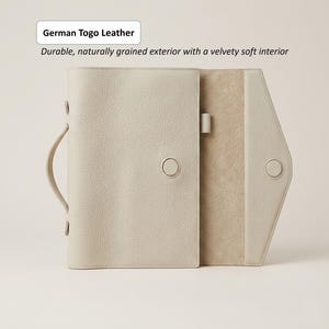 May include: A light beige leather journal with a handle and button closure. The journal is open, revealing a soft interior. The text on the image reads "German Togo Leather" and "Durable, naturally grained exterior with a velvety soft interior."