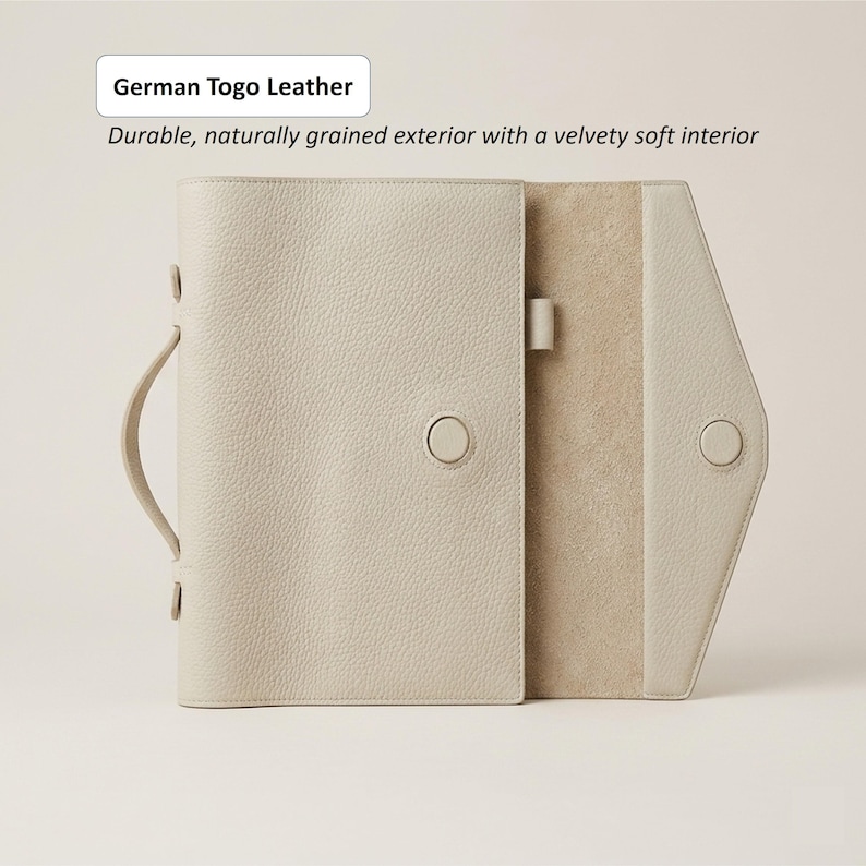 May include: A light beige leather journal with a handle and button closure. The journal is open, revealing a soft interior. The text on the image reads "German Togo Leather" and "Durable, naturally grained exterior with a velvety soft interior."