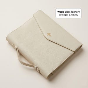 May include: Cream-colored leather portfolio with a flap closure and a gold cross emblem. Features a handle and a textured surface. The text "World-Class Tannery Perlinger, Germany" is visible.