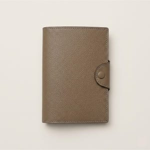 May include: A taupe-colored leather wallet with a button closure. The wallet has a textured surface and is rectangular in shape. The button is circular and matches the color of the wallet.