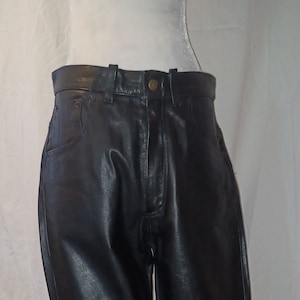 Puede incluir: Pantalón de cuero negro de pierna recta. El pantalón tiene una cremallera y trabillas para el cinturón.