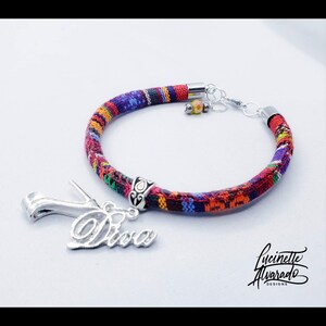 May include: A colorful fabric bracelet with a silver charm that says "Diva." The bracelet features a mix of red, blue, yellow, and purple patterns. The bracelet has silver end caps and a clasp. A small charm with yellow and orange beads is also attached.