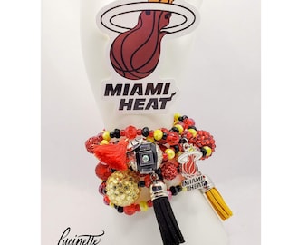 Może przedstawiać: Wielowątkowa bransoletka z czerwonymi, czarnymi i żółtymi koralikami, zawieszkami i frędzlami. Bransoletka posiada zawieszkę z logo Miami Heat oraz naklejkę z logo i nazwą drużyny. Prezentowana jest na białym, ręcznie ukształtowanym stojaku.