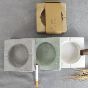 May include: Three concrete ashtrays with different colored interiors: gray, green, and white. A brown cigar rests on the white ashtray. A single cigarette and a brown paper box with the letters "L/R" are also in the image.