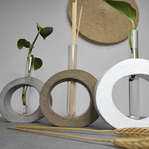 Puede incluir: Tres jarrones circulares de hormigón con tubos de vidrio para contener flores o vegetación. Los jarrones son de diferentes tonos de gris y blanco. Los jarrones están sobre una superficie gris con tallos de trigo secos.