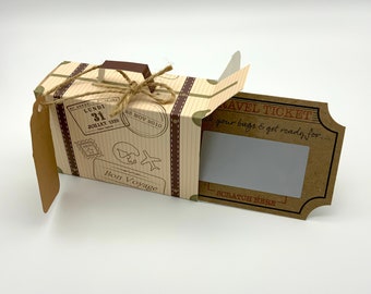 May include: A brown cardboard box shaped like a vintage suitcase with a brown paper travel ticket attached. The box has a brown handle, brown straps, and a brown paper tag. The travel ticket has the text "TRAVEL TICKET" and "Pack your bags & get ready for..." The box has the text "Bon Voyage" and a date stamp that says "LUNDI 31 JULLET 1939" and "23 NOV 2010".