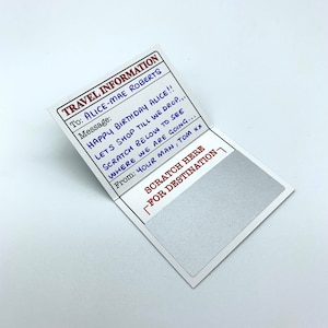 May include: A white card with red and black text that reads "TRAVEL INFORMATION" and "TO: ALICE-MAE ROBERTS". The card has a message that reads "HAPPY BIRTHDAY ALICE!! LETS SHOP TILL WE DROP... SCRATCH BELOW TO SEE WHERE WE ARE GOING... From: YOUR MAN, TOM XX". The card has a silver scratch-off area that says "SCRATCH HERE FOR DESTINATION".