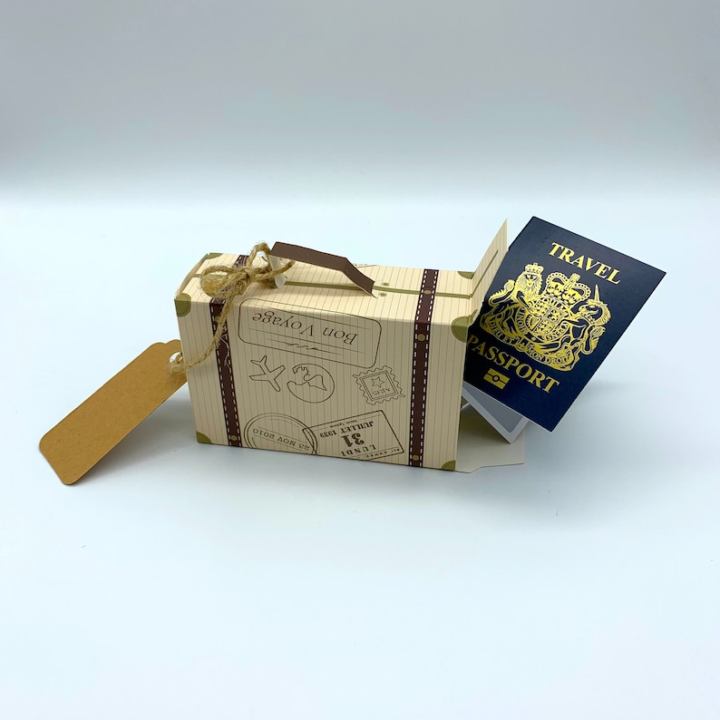 May include: A small cardboard suitcase with a brown handle and a tag. The suitcase is decorated with a travel theme, including a world map, a plane, and the words "Bon Voyage". A blue passport with the words "Travel Passport" is partially visible inside the suitcase.