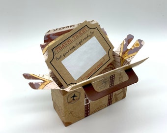 May include: A brown cardboard box shaped like a suitcase with a travel ticket attached to the top. The ticket says "Travel Ticket" and "Pack your bags & get ready for..." with a blank space to scratch off. The box has a brown handle and a small airplane graphic on the side.