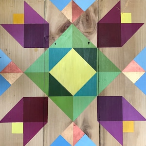 May include: A geometric quilt block with a central diamond shape in shades of green, blue, and yellow. The diamond is surrounded by four purple and yellow triangles, all on a wood background.
