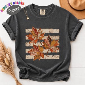 May include: A charcoal gray short-sleeve t-shirt with a crew neckline. The front features a graphic of three maple leaves with various patchwork patterns in orange, brown, and cream colors, layered over horizontal distressed white stripes.