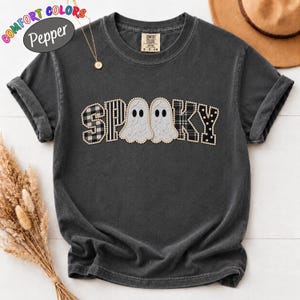 May include: A charcoal grey short-sleeve t-shirt featuring the word SPOOKY in a decorative font. The letters S and P have a black and white plaid pattern, while the O letters are replaced by two white ghost illustrations. The K and Y have a black and white polka dot pattern.