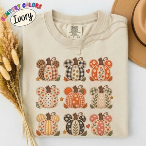 以下が含まれることがあります： アイボリー色のTシャツで、ネズミの耳の形をした9つのカボチャのイラストがグリッド状に配置されています。カボチャには、チェック柄、水玉模様、花柄、アニマルプリントなど、オレンジ、ブラウン、クリーム色の様々なパターンが描かれています。
