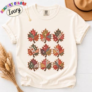 May include: An ivory short-sleeve t-shirt featuring a graphic print of nine maple leaves arranged in a grid. Each leaf displays a unique patchwork design with various patterns including plaid, floral, and polka dots in red, green, and orange tones.