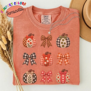 May include: A terracotta-coloured crew neck t-shirt featuring a graphic print of three pumpkins and three bows arranged in a grid. The pumpkins display floral and checkered patterns, while the bows feature polka dot and plaid designs.
