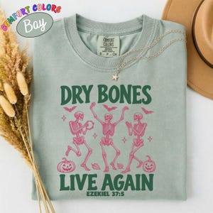 May include: A sage green t-shirt featuring a graphic of three pink skeletons dancing with pumpkins, bats, and stars. The text reads "DRY BONES LIVE AGAIN EZEKIEL 37:5" in dark green lettering. The shirt has a crew neckline and a Comfort Colors brand label.