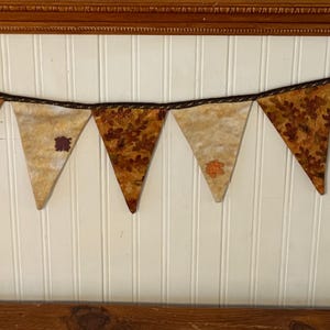 May include: A decorative banner with seven triangular flags. The flags are in shades of tan and brown, with a patterned design. The banner is strung on a dark brown ribbon with dark brown ribbons at each end. The banner is hanging on a white wall.