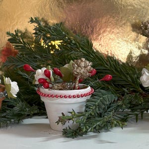 Könnte beinhalten: Drei Miniatur-Topfpflanzen, die mit roten und weißen Weihnachtsornamenten verziert sind. Die Töpfe sind mit Strasssteinen, roten Perlen und weißen Perlen verziert.