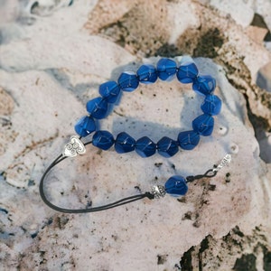 May include: A blue gemstone mala with a silver charm. The mala is made of faceted beads and is strung on a black cord.