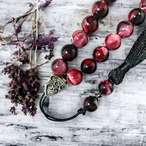 Könnte beinhalten: Eine rote und schwarze Edelstein-Mala mit einem silbernen Anhänger. Die Mala besteht aus runden Perlen und hat einen schwarzen Quasten.