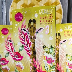 May include: Three boxes of Aurifil thread, each featuring a colorful illustration of a giraffe in a jungle setting. The boxes are labeled "Neons & Neutrals Thread Collection" and feature the Aurifil logo. Each box contains 3 new colors.