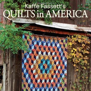 May include: A colorful quilt with a geometric pattern of cubes. The quilt is made of fabric with a variety of colors and patterns. The quilt is hanging on a wooden wall with vines growing on it. The text "Kaffe Fassett's QUILTS in AMERICA" is at the top of the image. The text "DESIGNS INSPIRED BY VINTAGE QUILTS FROM THE AMERICAN MUSEUM IN BRITAIN" is at the bottom of the image.