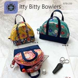 May include: A sewing pattern for two sizes of small, fabric bags with a top handle. The bags are shown in different floral prints with a navy blue trim. The pattern is called "Itty Bitty Bowlers" and the pattern number is ATB-233. The text "The cutest way to keep track of your little stuff. Fat Quarter Friendly. Two sizes included." is at the bottom of the image.