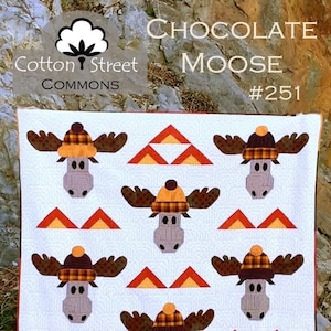 Op de afbeelding: Een wit quilt met een patroon van bruine elanden die geruite hoeden en oranje driehoeken dragen. De quilt is getiteld "Chocolate Moose #251" en is een ontwerp van Cotton Street Commons.