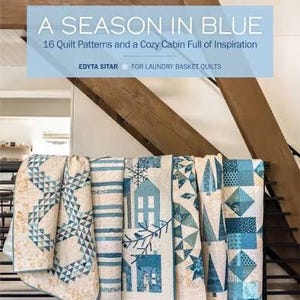 May include: A display of blue and white quilts, showcasing various patterns, including geometric shapes and house motifs. The quilts are draped over a black metal rack. The image also features a book cover with the title "A Season in Blue".