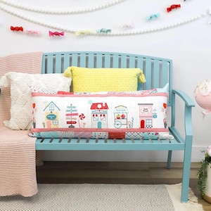 Puede incluir: Un cojín rectangular con un diseño de pueblo caprichoso con edificios coloridos, una tienda de golosinas y letreros. El cojín descansa sobre un banco azul con otras almohadas y una manta rosa. Una guirnalda de perlas y decoraciones de caramelos cuelgan encima.