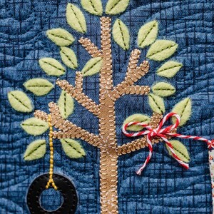 May include: A close-up of a blue fabric with a brown and green embroidered tree. The tree has a tire swing and a red and white ribbon tied to a branch. The fabric has a grid pattern and the text "KIMBERBELL" is visible at the bottom.