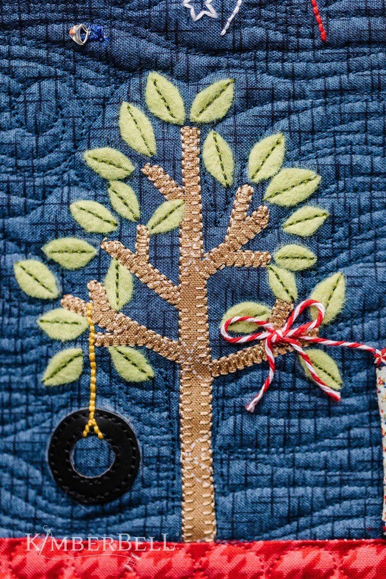 May include: A close-up of a blue fabric with a brown and green embroidered tree. The tree has a tire swing and a red and white ribbon tied to a branch. The fabric has a grid pattern and the text "KIMBERBELL" is visible at the bottom.
