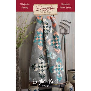 May include: A gray quilt with a repeating pattern of colorful squares and the text "English Knot." The quilt measures 68" x 91". The quilt is hanging on a wooden wall with a pitchfork and vase in the background. The quilt is part of the "Stashtastic Pattern Series!"
