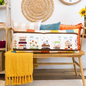 May include: A long rectangular quilted pillow featuring a fall-themed scene with a general store, sunflowers, a truck carrying pumpkins, a cider stand, and a house. The pillow is accented with brown borders and sits on a wooden bench with a yellow throw.
