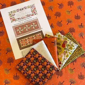 Op de afbeelding: Een quiltset voor een loper genaamd "The Orchard" met een patroon van bomen en eekhoorns. De set bevat stofvierkanten in tinten oranje, groen, bruin en wit. De stofvierkanten zijn gerangschikt op een achtergrond van oranje stof met witte stippen.