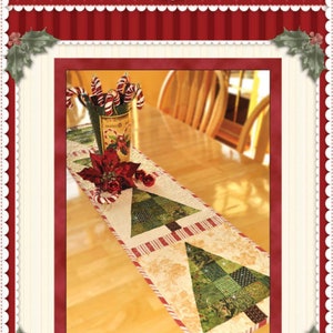 May include: A patchwork Christmas tree table runner with two green trees on a cream background. The runner has red and white stripes and a gold border. The runner is on a wooden table with a vase of candy canes and a poinsettia.