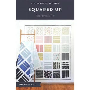 May include: A quilt with a grid pattern of various fabrics in pastel colors, including pink, yellow, and blue, is displayed on a wall. A second quilt with a geometric design in blue, gray, and black hangs on a wooden ladder. The text "SQUARED UP" is at the top.