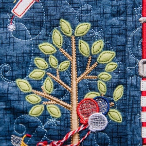 May include: A blue and red embroidered fabric with a tree, a bicycle, and balloons. The tree has green leaves and a brown trunk. The bicycle is yellow with a basket and red ribbons. The balloons are red, white, and blue.