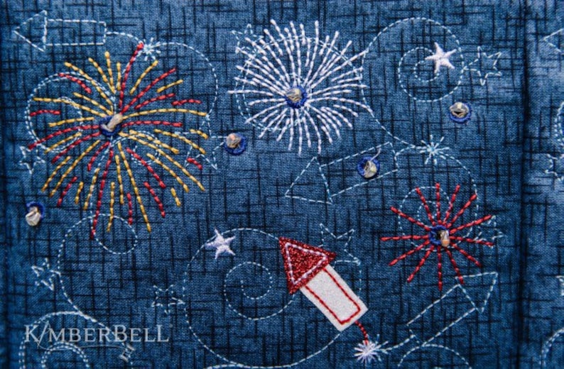 May include: Embroidered fabric with a blue background and white, red, and yellow fireworks designs. The fabric also features a red and white rocket design and the text "KIMBERBELL".