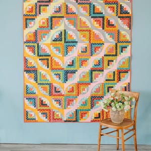 May include: A colorful quilt with a geometric pattern of squares and curved shapes in shades of yellow, orange, blue, and pink. The quilt is hanging on a light blue wall. A wooden chair with a woven basket of flowers sits below.