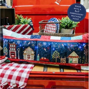 May include: A red, white, and blue embroidered bench pillow with a scene of a small town with fireworks. The pillow is made with a 5x7 embroidery machine hoop. The pillow is sitting on the back of a red pickup truck. The text "Main Street Celebration Bench Pillow" is at the bottom of the image. The text "Formats: ART, DST, EXP, HUS, JEF, PES, VIP, VP3, XXX" is below the title.